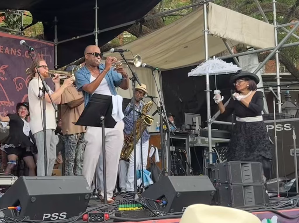 Musician and Trumpeter James Andrews and fellow musicians performing on stage in the French Quarter Festival. Musicians are playing the trumpet and saxophone in casual dress performing music on a stage. 